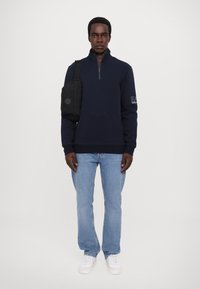 Sudadera de forro polar azul marino con cremallera y dobladillo acanalado, con un logo detallado en la manga, combinada con unos vaqueros de mezclilla clara y zapatillas blancas.