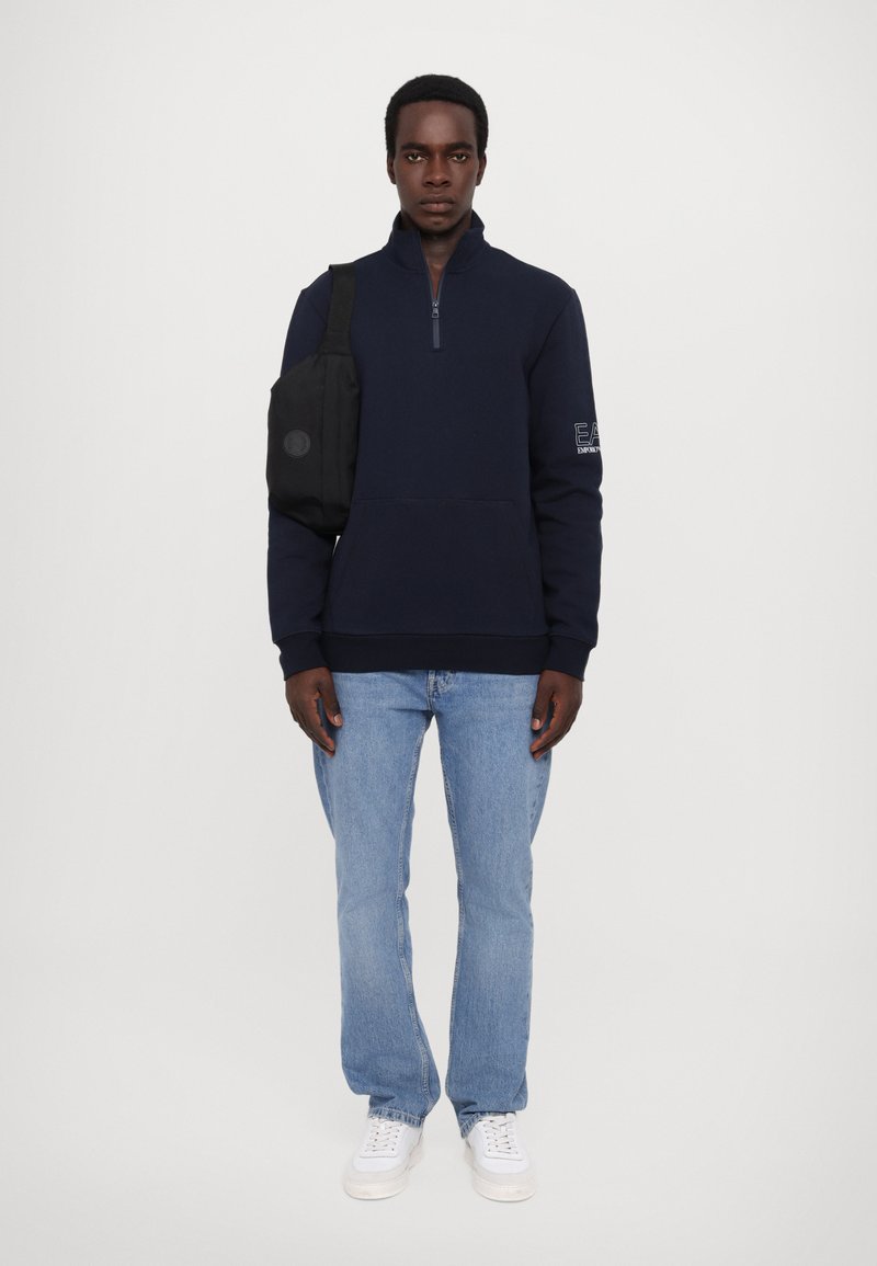 Sudadera de forro polar azul marino con cremallera y dobladillo acanalado, con un logo detallado en la manga, combinada con unos vaqueros de mezclilla clara y zapatillas blancas.