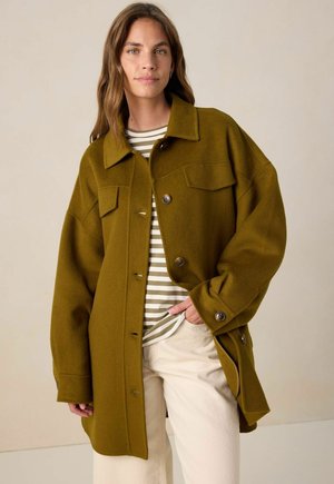 Olive green wool jacket with a button-up front, large chest pockets, and a relaxed fit. Paired with a striped shirt underneath.