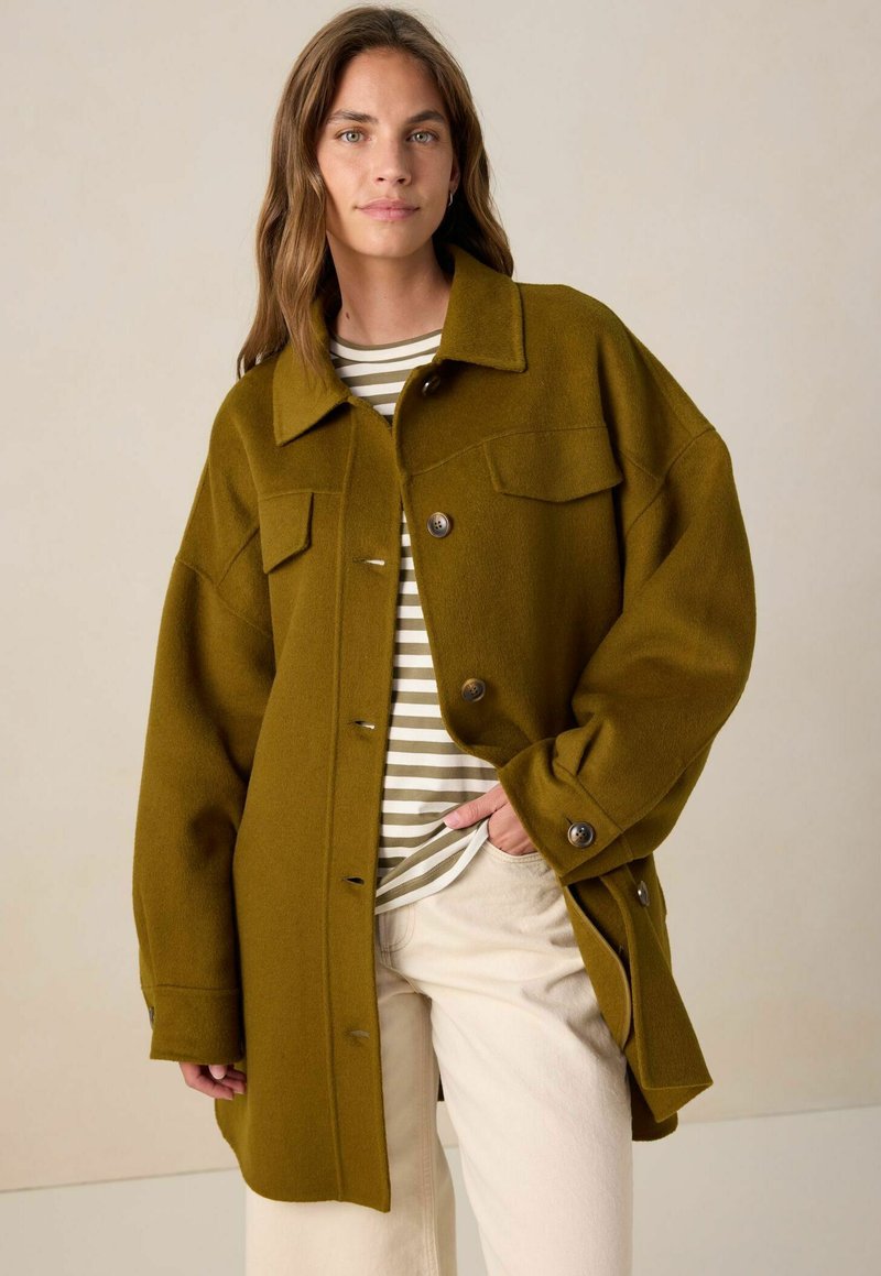 Olivgrüne Wolljacke mit einer Knopfleiste, großen Brusttaschen und einem lockeren Schnitt. Kombiniert mit einem gestreiften Hemd darunter.