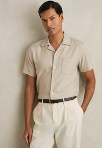 Camisa beige de manga corta con cuello, un bolsillo en el pecho y botones, combinada con pantalones crema y un cinturón oscuro.