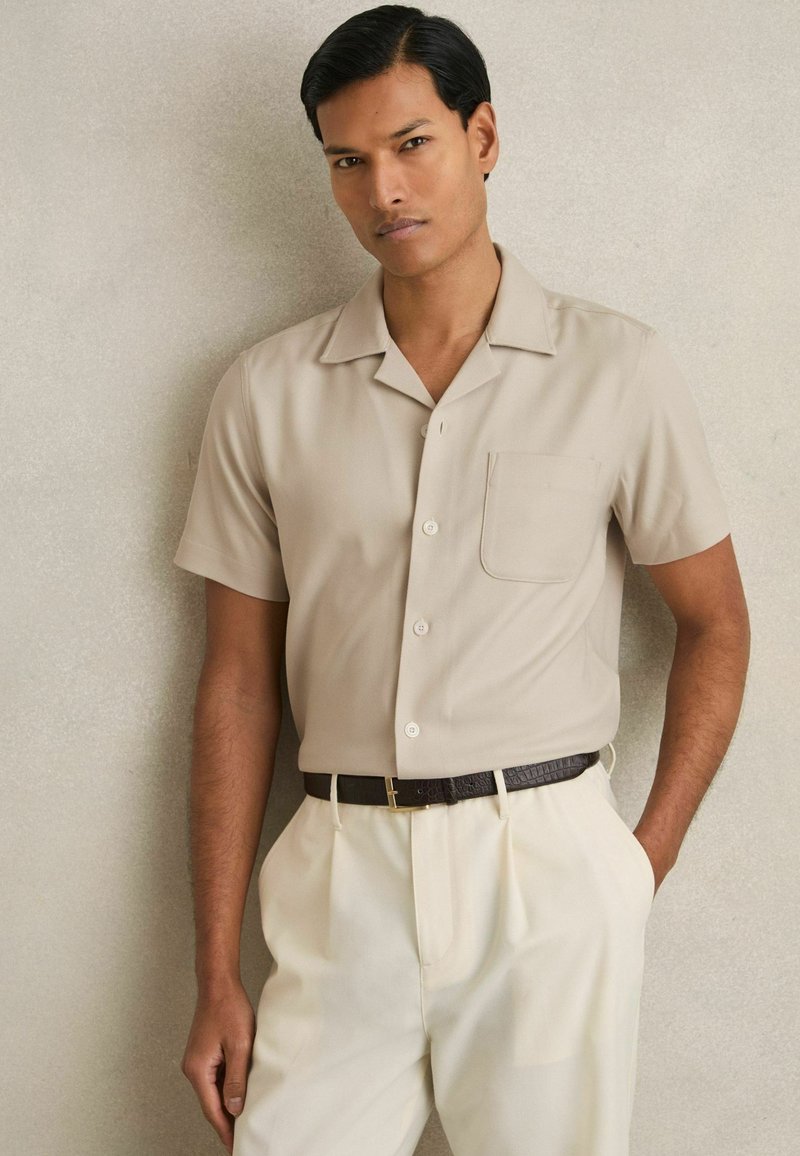 Beige Kurzarmhemd mit Kragen, einer Brusttasche und Knöpfen, kombiniert mit cremefarbenen Hosen und einem dunklen Gürtel.