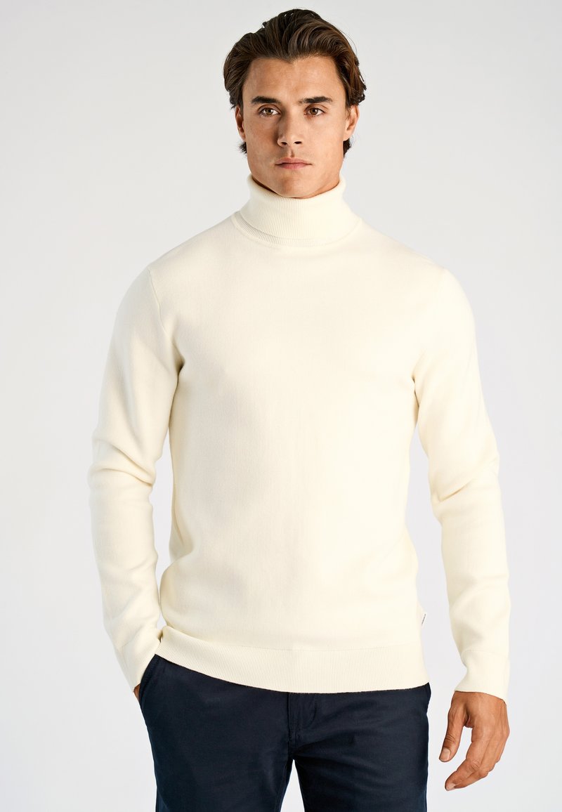 Helles, cremefarbenes Rollkragenpullover aus weichem Stoff, tailliertes Design, gerippte Bündchen und Saum, getragen mit dunkler Hose.