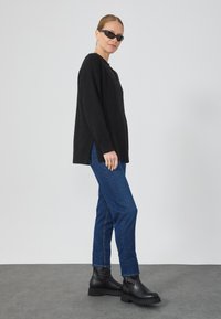 Sweater negro de gran tamaño con aberturas laterales, combinado con jeans rectos azules y botines negros de suela gruesa. La modelo lleva gafas de sol negras.