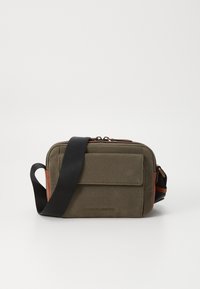 Sac bandoulière en toile vert olive avec une poche avant, des accents marron, une sangle noire, et une fermeture éclair. Présente le logo sur le devant.