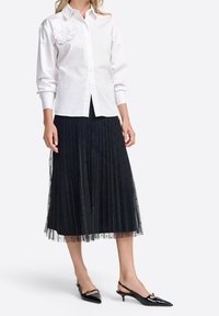 Weiße Bluse mit floralem Akzent, kombiniert mit einem schwarzen, plissierten Tüllrock mit Polka-Dots und schwarzen, spitz zulaufenden Slingback-Pumps.