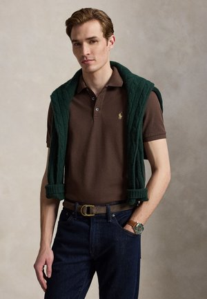 Jeune homme portant un polo marron, un jean foncé, une montre et un pull vert drapé sur ses épaules, avec un fond uni.