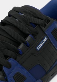 Gros plan sur une chaussure Globe noire et bleue avec de grosses lacets noirs et des détails en tissu mesh rembourré.