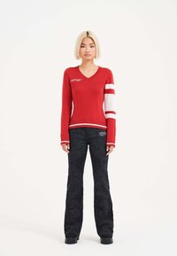Roter gestrickter Pullover mit V-Ausschnitt und weißen Streifen auf einem Ärmel, kombiniert mit schwarzen Hosen mit geprägten Spiralmustern.