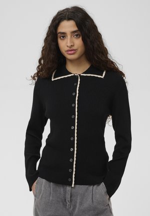Jonge vrouw met krullend haar die een zwarte geribbelde trui met knopen draagt met witte bies langs de kraag en voorkant, handen in de zakken van grijze broek.