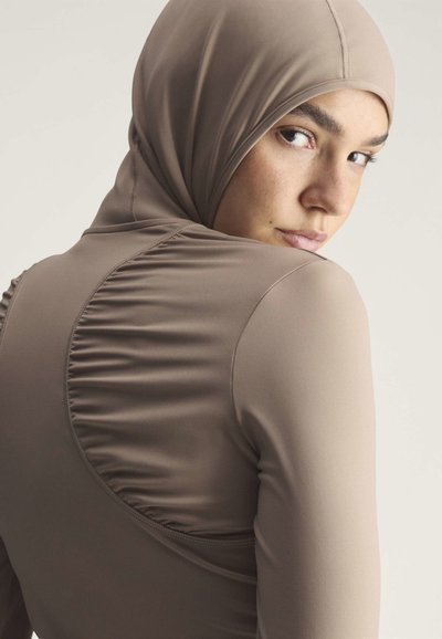 Young woman looking over her shoulder wearing a fitted taupe hooded top with ruched detailing on the back against a neutral background.