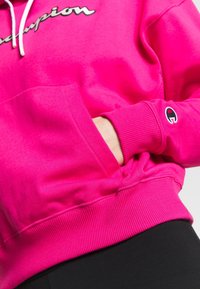 Sweat à capuche Champion rose vif avec cordons blancs, une main dans la poche avant et le logo Champion sur la poitrine et le bord de la manche.