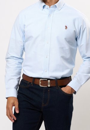 Camisa azul claro de manga larga abotonada al frente, combinada con jeans oscuros de mezclilla y un cinturón de cuero marrón con hebilla plateada.