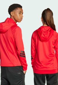 Sudaderas deportivas rojas con acentos de franjas negras en las mangas. Tejido suave y que absorbe la humedad. Vista trasera; combinadas con pantalones deportivos negros.