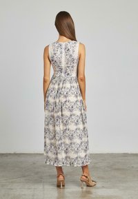 Maxikleid in hellem Creme mit lila Blumenmuster. Verfügt über ein tailliertes Oberteil, einen gefalteten Rücken und einen gewellten Saum. Kombiniert mit neutralen Absätzen.