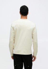 Κρέμ πουκάμισο μακρυμάνικο με στρογγυλή λαιμόκοψη και απαλό ύφασμα. Διαθέτει απλό σχέδιο· φοριέται με μαύρο παντελόνι, και φαίνεται από πίσω.