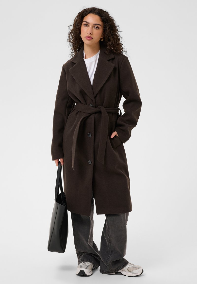 Femme portant un manteau ceinturé marron foncé, un pantalon large gris, des baskets blanches, tenant un sac fourre-tout noir sur un fond clair uni.