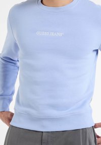 Světle modrý svetr s žebrovanými manžetami a lemem, s bílou vyšívanou logem "GUESS JEANS" na hrudi, vyrobený z měkkého materiálu.