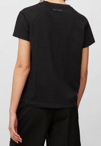 Persona que lleva una camiseta negra de manga corta y pantalones negros, mostrada de espaldas con el texto "Marc O'Polo" cerca del cuello.