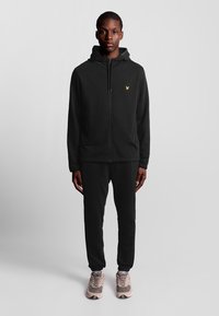 Felpa nera con zip e logo giallo sul petto, abbinata a pantaloni jogger neri e sneakers beige. Il tessuto sembra morbido e liscio.