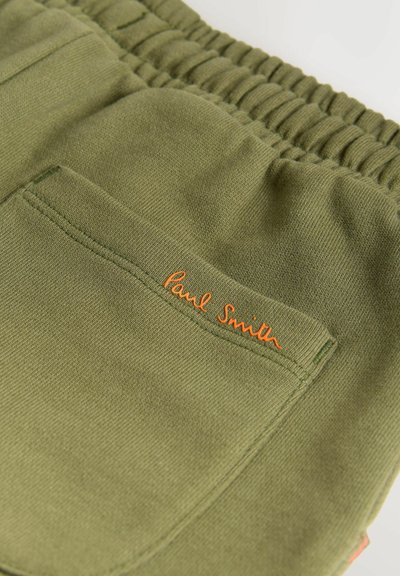 Nærbillede af olivengrønt stof med elastisk taljebånd og en baglomme broderet med "Paul Smith" i orange tråd.