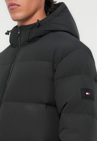 Zwarte pufferjack met een capuchon, voorzien van gestikte secties, een ritssluiting en een logopatch op de linkermouw. Soepele, matte textuur.