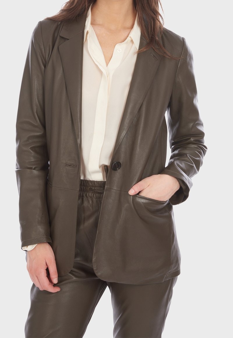 Femme portant un blazer en cuir marron et un pantalon assorti sur une chemise blanche boutonnée, debout avec une main dans la poche.