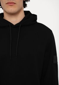 Felpa nera con cappuccio, dotata di cordino, caratterizzata da una texture morbida e un design minimalista. Include una toppa sul maniche per un tocco di dettaglio in più.