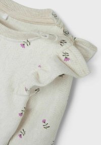 Cremefarbiges Kleidungsstück mit Fischgrätmuster, das kleine lila Blumenmuster und einen Rüschensaum mit Druckknöpfen aufweist. Weiche Textur.