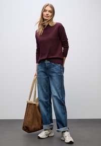 Burgunderfarbener Strickpullover, lockere blaue Jeans mit Seitenstreifen und eine braune Wildledertasche. Graue Sneakers mit hellen Akzenten. Minimalistischer Hintergrund.