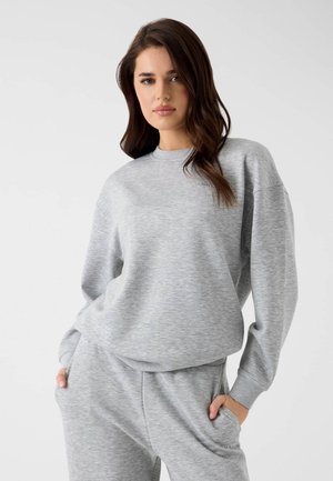 Junge Frau trägt ein hellgraues Sweatshirt und passende Jogginghose, steht mit den Händen in den Taschen vor einem schlichten Hintergrund.