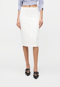 Γυναίκα που φοράει άσπρη pencil φούστα μέχρι το γόνατο και μαύρες mule γόβες με μυτερή μύτη και λουράκια με αγκράφες, στέκεται μπροστά σε απλό φόντο.