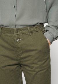 Pantalon en coton vert olive avec fermeture à boutons, poches latérales et une discrète étiquette logo, associé à un chemisier gris clair.