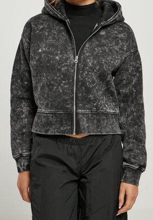 Frau trägt einen schwarzen Acid-Wash-Kapuzenpullover mit Reißverschluss über einem schwarzen Rollkragenpullover und schwarzen strukturierten Hosen, steht vor einem schlichten Hintergrund.