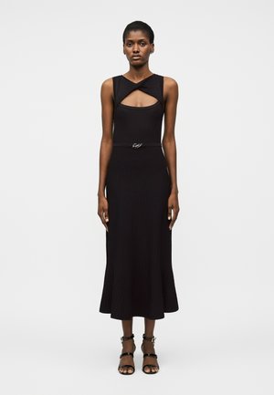 Kobieta w czarnej, bezrękawnikowej sukience midi z skręconym wycięciem przy dekolcie oraz czarnych sandałach na wysokim obcasie z paskami, stojąca na białym tle.