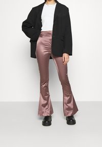 Czarna oversize marynarka, biały krótki top i różowe satynowe spodnie z szerokimi nogawkami. Buty to czarne, masywne botki na obcasie. Minimalistyczne tło.