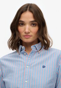 Kvinde med skulderlangt brunt hår iført en blå skjorte med lyserøde lodrette striber og et lille navy-logo på brystet.