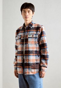 Camicia di flanella a quadri blu, arancione e nera, con chiusura a bottoni frontale, due tasche sul petto e un'etichetta di lato. Texture morbida.