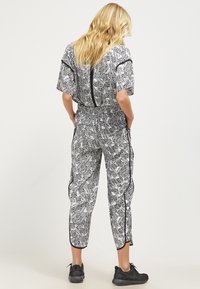 Femme aux cheveux blonds portant une combinaison à motifs de feuilles noir et blanc avec des manches courtes et des baskets noires, se tenant les mains dans les poches.