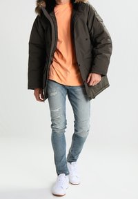 Giacca puffer verde oliva con cappuccio foderato in pelliccia, abbinata a una t-shirt pesca e jeans chiari strappati. Sneakers bianche completano il look.
