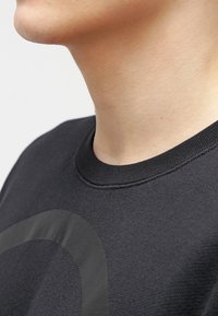 Svart sweatshirt med rund hals, texturerad tyg och en stor, subtil grafisk design på framsidan.