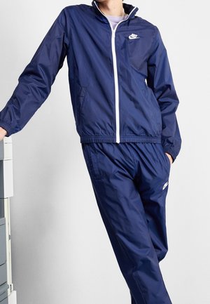 Osoba ubrana w granatowy dres Nike z białym zamkiem i logo, stojąca z jedną ręką w kieszeni, a drugą opartą na białej powierzchni.