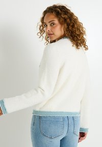 Suéter blanco suave con un ribete azul claro en los puños y el dobladillo, que presenta un corte holgado y una textura suave. Llevado con jeans azules.