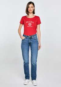 Červené bavlněné tričko s grafikou "HOLLY JOLLY" a bílým písmem, spárované s modrými džíny a bílými teniskami.