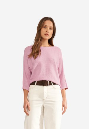 Femme aux longs cheveux bruns portant un pull côtelé violet clair, un pantalon blanc et une ceinture sombre, debout devant un fond blanc.