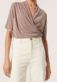 En wrap-stil, kortärmad topp i taupe-färgad tyg med mjuka draperingar. Kombinerad med högmidjade, vita denimjeans med knäppning.