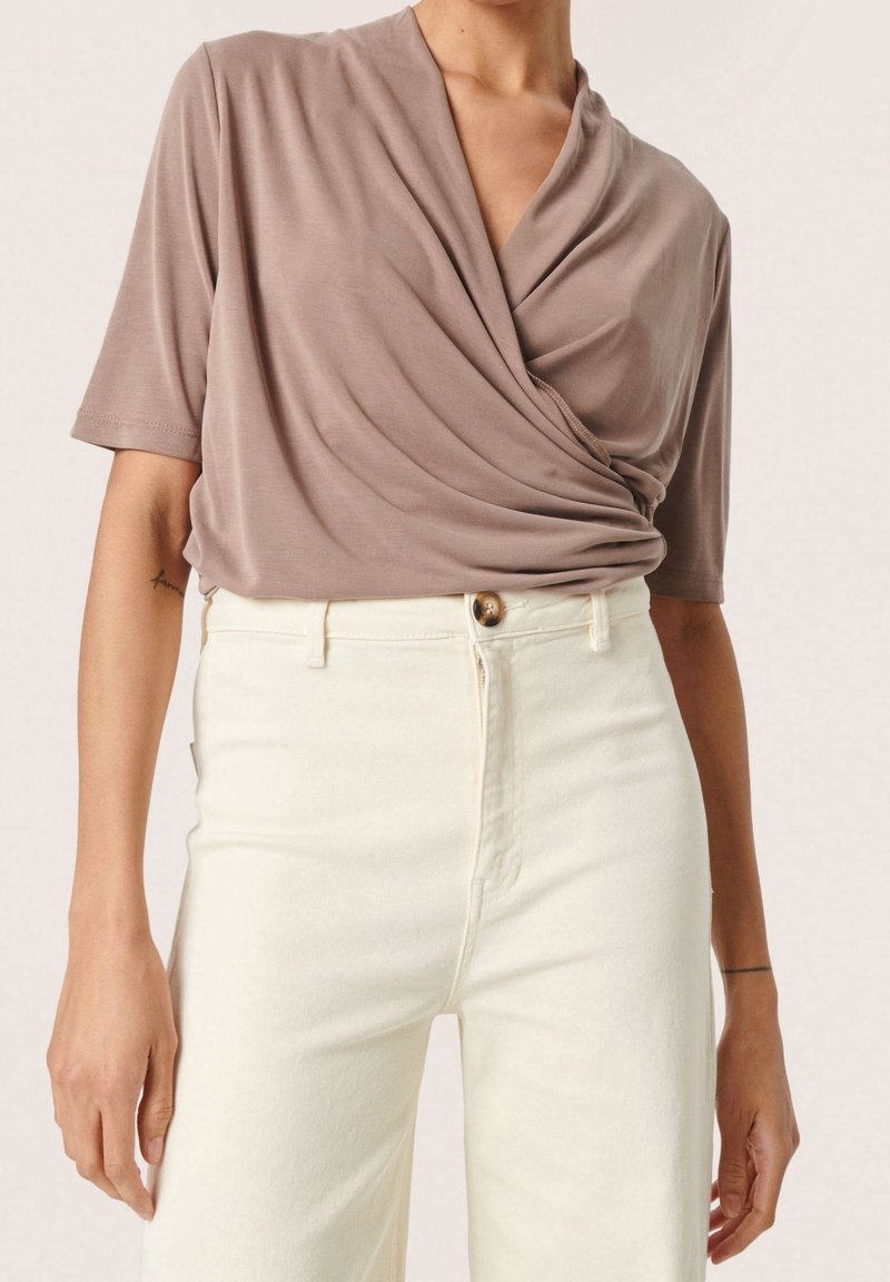 En wrap-stil, kortärmad topp i taupe-färgad tyg med mjuka draperingar. Kombinerad med högmidjade, vita denimjeans med knäppning.