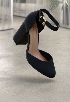 Black high-heeled shoe with a smooth texture, pointed toe, and a thick block heel. Features an ankle strap with a gold-tone buckle.