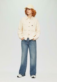 Beige corduroy overhemd met knoopsluiting, twee borstzakken; lichtblauwe wijde jeans; crèmekleurige cap; zwart-witte sneakers.