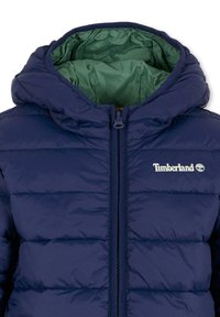 Giacca imbottita blu navy con chiusura a zip, grande cappuccio e fodera interna verde a contrasto. Presenta il logo Timberland sul petto.